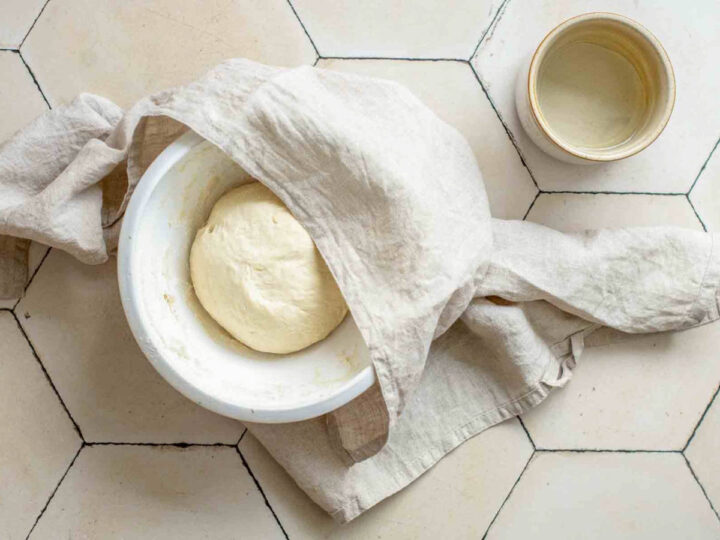 A white bowl containing raw dough sits on a hexagonal tiled surface, partially covered by a light-colored linen cloth. A small ceramic cup of water is nearby.