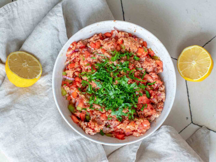 A white bowl filled with a mixture of minced meat, diced tomatoes, red onions, and green peppers, topped with a generous handful of fresh chopped parsley. The bowl sits on a light-colored cloth with two lemon halves placed on either side.