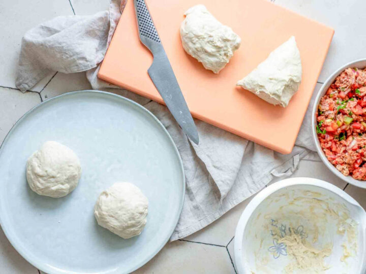 An overhead view shows the process of portioning dough for lahmacun on a white hexagonal tiled floor. Two smooth, round dough balls rest on a light blue plate, while two irregular pieces of dough and a metal kitchen knife sit on an orange cutting board. In the surrounding area, there is an empty white mixing bowl and a separate bowl containing the prepared meat and vegetable topping.
