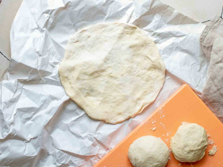 A single dough ball rolled into a thin, flat circle on crumpled white parchment paper, with more dough balls visible on an orange board nearby.