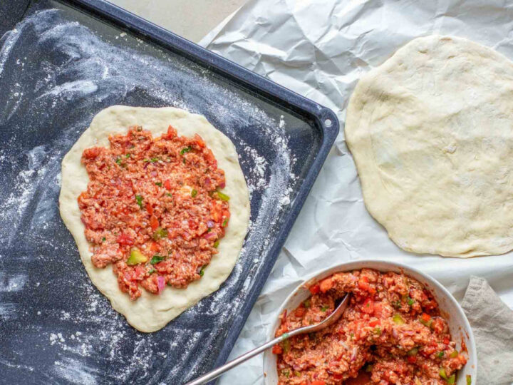A baking sheet sprinkled with flour holds one thin circle of raw dough, which is topped with an even layer of the meat and vegetable mixture. Beside the tray, a second plain dough round rests on white parchment paper, and a bowl of the remaining topping with a spoon is visible in the foreground.