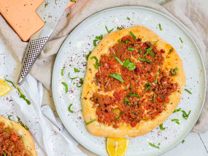 A freshly baked, golden-brown Turkish lahmacun served on a light blue plate, topped with a savory meat and vegetable mixture and garnished with fresh parsley. The plate is dusted with red sumac spice and accompanied by a lemon wedge on a rustic tabletop.