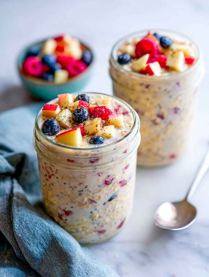 Two glass jars of apple berry overnight oats topped with diced apples and mixed berries on a white marble surface with a cool-toned napkin and spoon nearby.