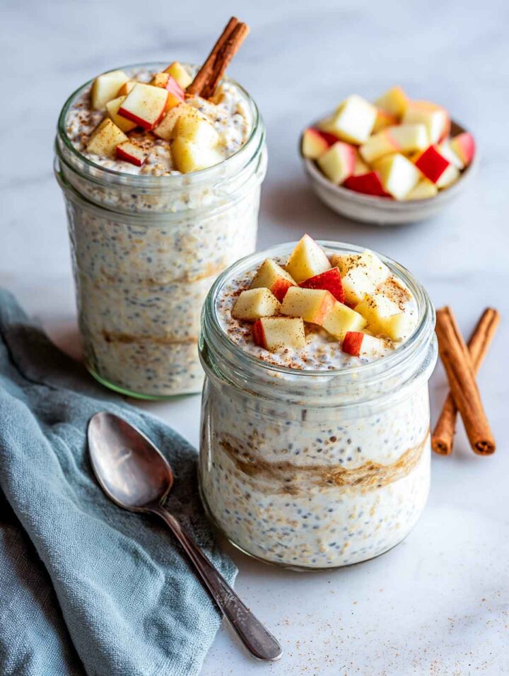 Two glass jars of apple cinnamon bran overnight oats topped with diced apples and cinnamon on a white marble surface with a soft sage napkin.