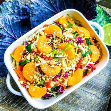 Colorful Asian cabbage salad in a white baking dish with mandarin orange segments, shredded cabbage, carrots, herbs, and chopped peanuts on a rustic table.
