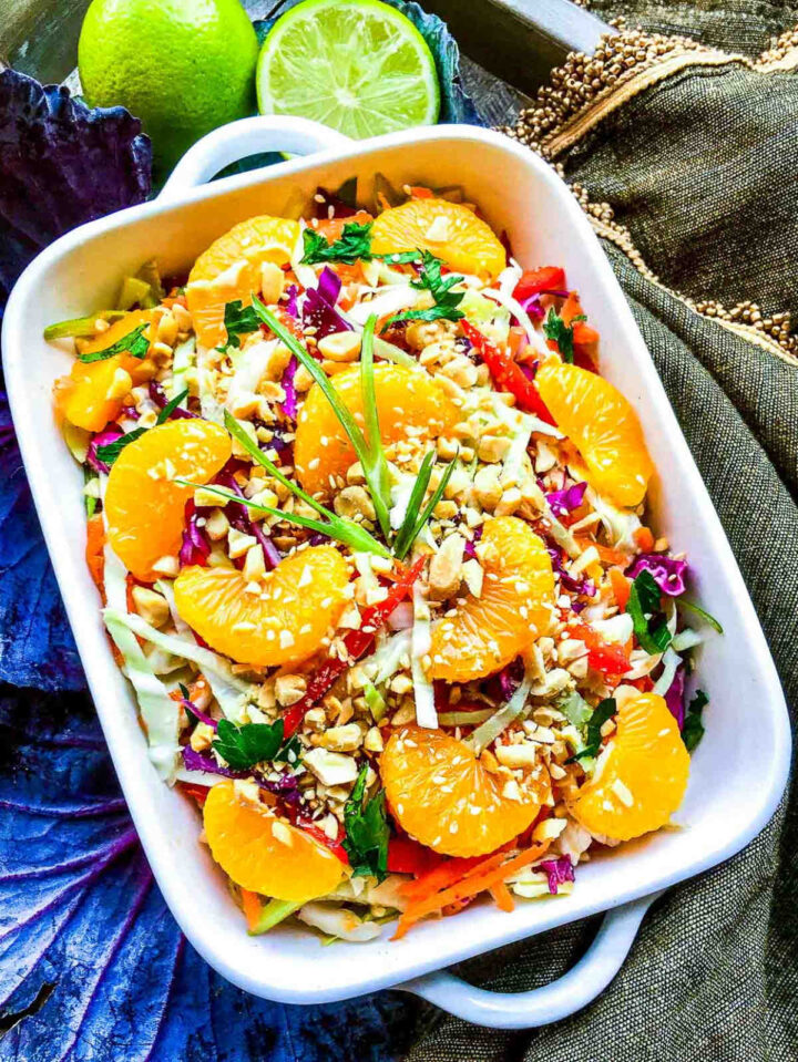 Asian cabbage salad topped with mandarin orange slices, chopped peanuts, herbs, and sesame seeds in a white dish, with lime and purple cabbage in the background.