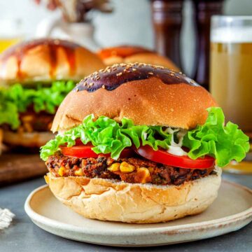Close-up of a black bean veggie burger on a sesame-topped bun with lettuce, tomato, and sauce, served on a plate with another burger and drinks in the background.