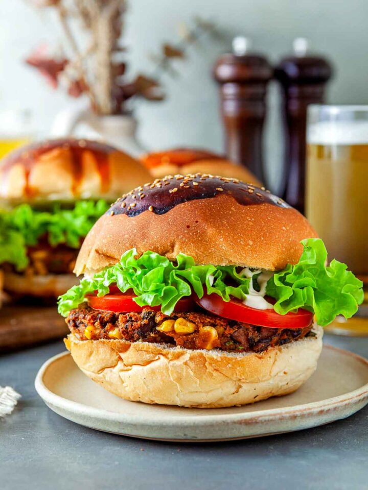 Close-up of a black bean veggie burger with a seeded bun, crisp lettuce, sliced tomato, and creamy sauce, plated with another burger and a glass of beer blurred in the background.