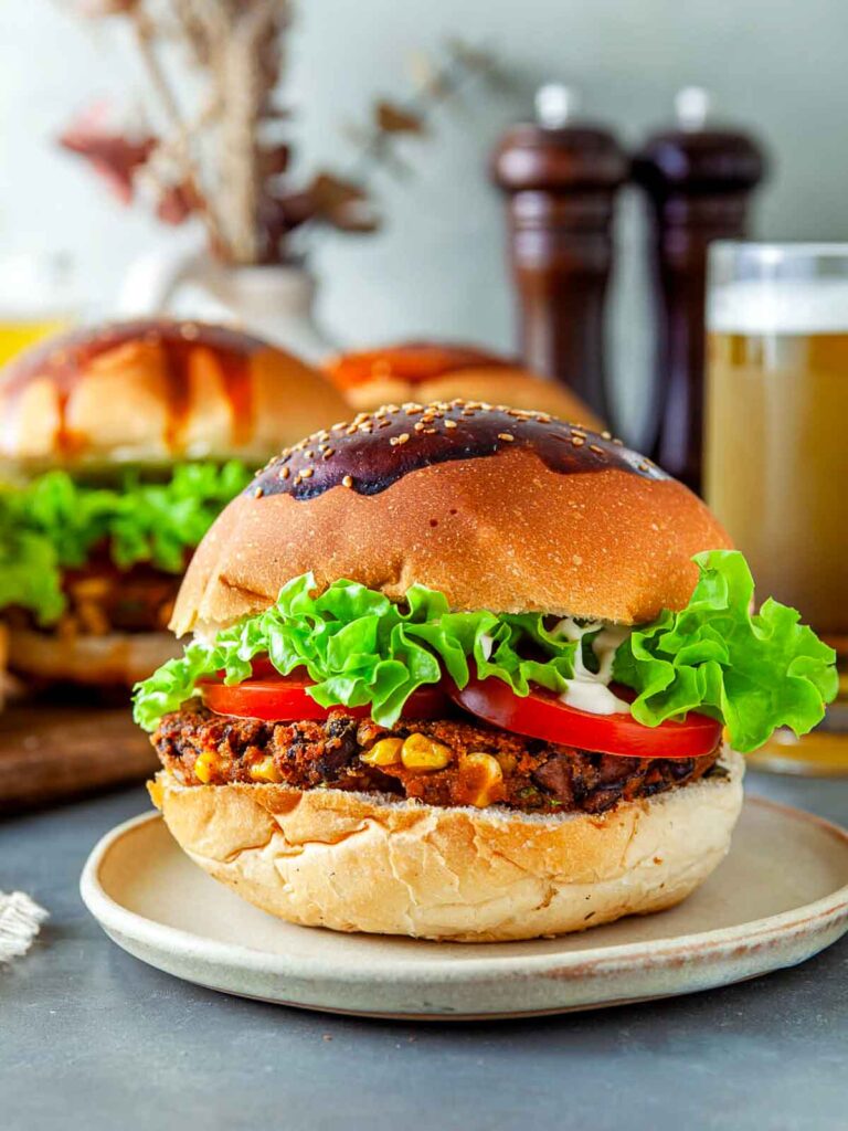 Two black bean veggie burgera with bun, crisp lettuce, sliced tomato, and creamy sauce, plated with another burger and a glass of beer blurred in the background.