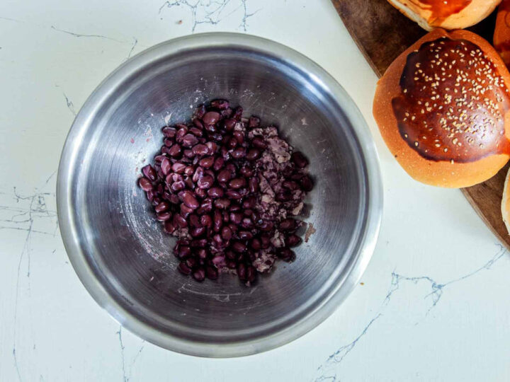 Overhead view of cooked black beans in a metal mixing bowl, partially mashed, with burger buns visible on the side.
