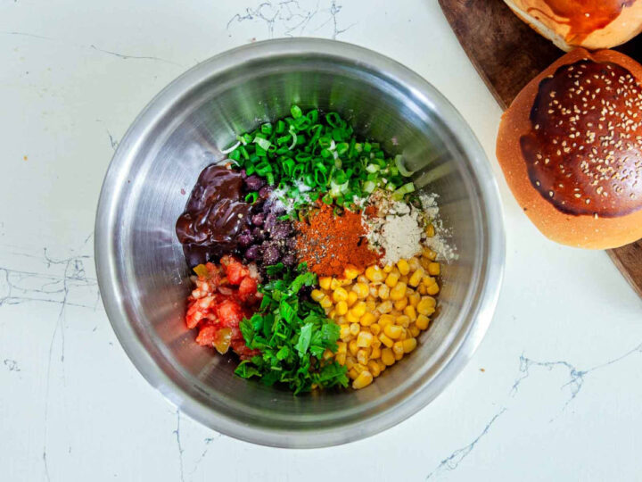 Overhead view of a metal mixing bowl filled with black beans, corn, chopped green onions, herbs, spices, and sauce, ready to be mixed for veggie burgers.