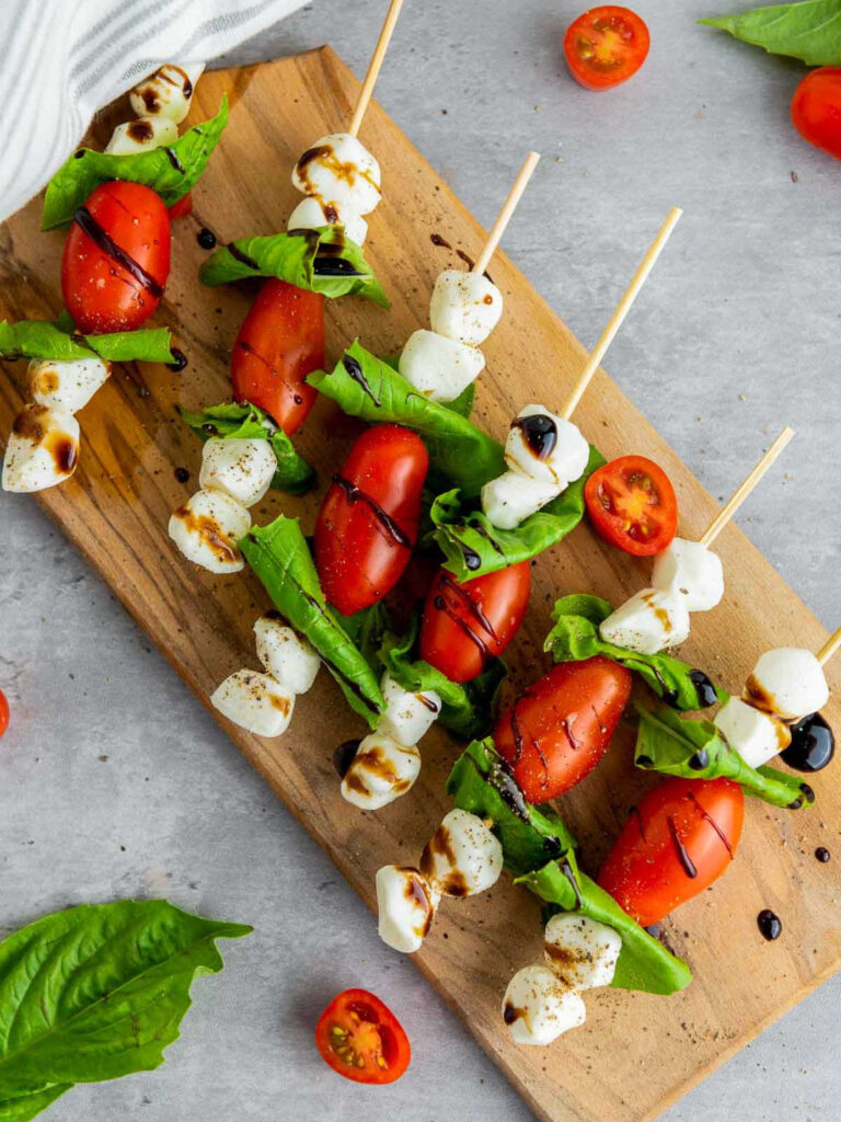 Caprese skewers with cherry tomatoes, fresh mozzarella, and basil leaves threaded on wooden sticks, drizzled with balsamic glaze and lightly seasoned, arranged on a wooden board.