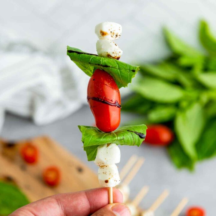 Hand holding a finished caprese skewer stacked with mozzarella pearls, folded basil leaves, and a cherry tomato drizzled with balsamic glaze, with a soft-focus background of fresh basil and additional skewers
