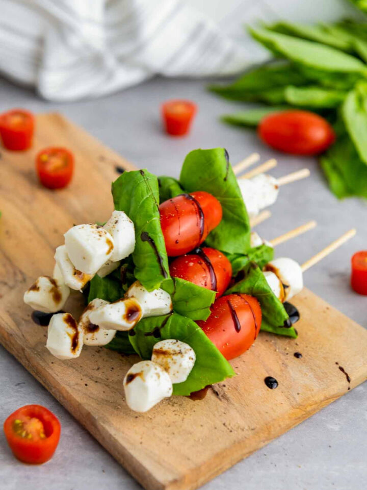 Caprese skewers stacked on a wooden board with cherry tomatoes, fresh mozzarella, and basil leaves, drizzled with balsamic glaze and lightly seasoned, with extra tomato pieces scattered around.