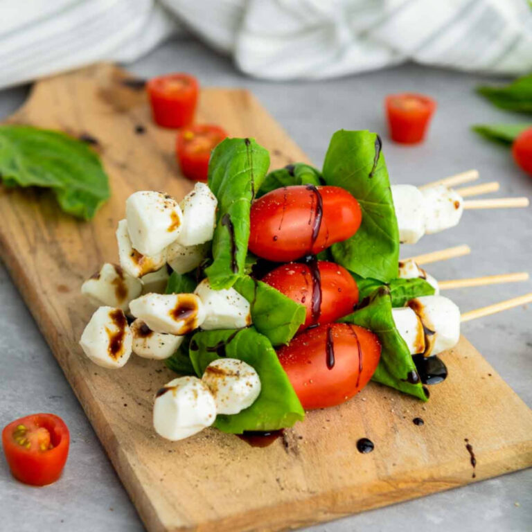 Caprese skewers stacked on a wooden board with cherry tomatoes, fresh basil leaves, and mozzarella pearls, lightly seasoned and drizzled with balsamic glaze, styled on a neutral kitchen surface with a soft linen backdrop.