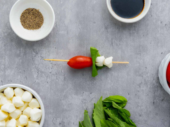 Single Caprese skewer assembled with a cherry tomato, fresh basil leaf, and mozzarella pearls on a wooden skewer, shown on a gray surface with bowls of pepper, balsamic glaze, basil, and mozzarella nearby.