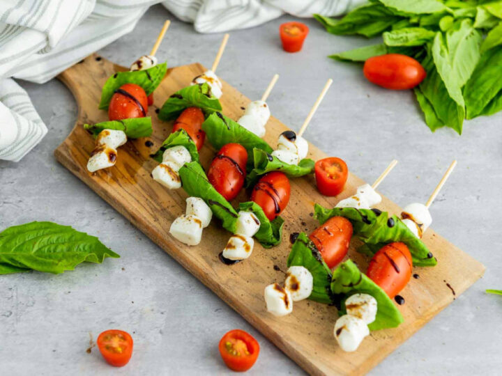 Caprese skewers arranged on a wooden serving board with cherry tomatoes, fresh basil leaves, and mozzarella pearls drizzled with balsamic glaze, with extra tomatoes and basil scattered on a light gray surface.