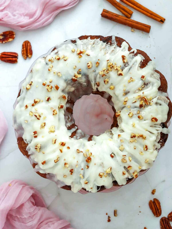 Overhead view of a whole carrot bundt cake topped with thick white glaze and chopped pecans, surrounded by cinnamon sticks and loose nuts on a marble surface.