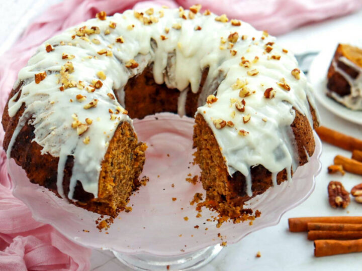 A carrot bundt cake on a glass stand with one slice removed, revealing the moist interior texture, topped with white frosting and pecans.
