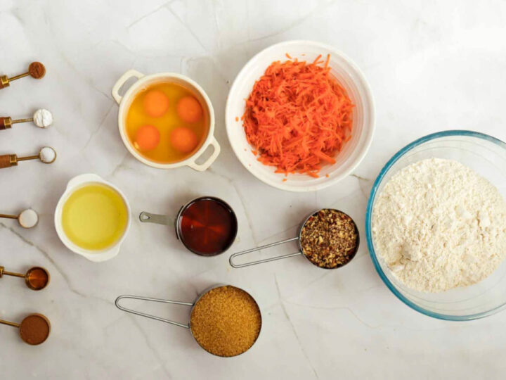 Flat lay of carrot cake ingredients including shredded carrots, eggs, flour, brown sugar, oil, chopped pecans, and various spices in measuring spoons.