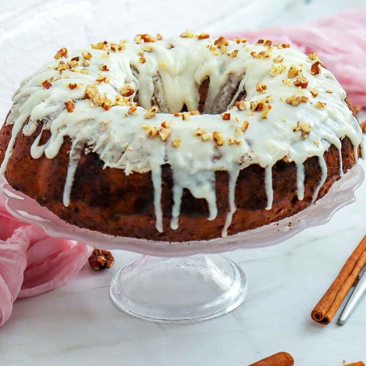 Side view of a glazed carrot bundt cake on a glass stand, showing thick white icing dripping down the sides and topped with crushed pecans.