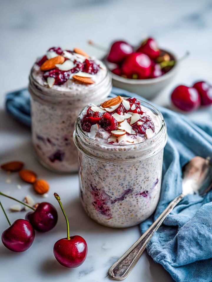 Two glass jars of cherry almond chia pudding with rolled rye, filled close to the rim and topped with chopped cherries and sliced almonds, styled on a white marble surface with fresh cherries, a blue-gray linen napkin, and a spoon.