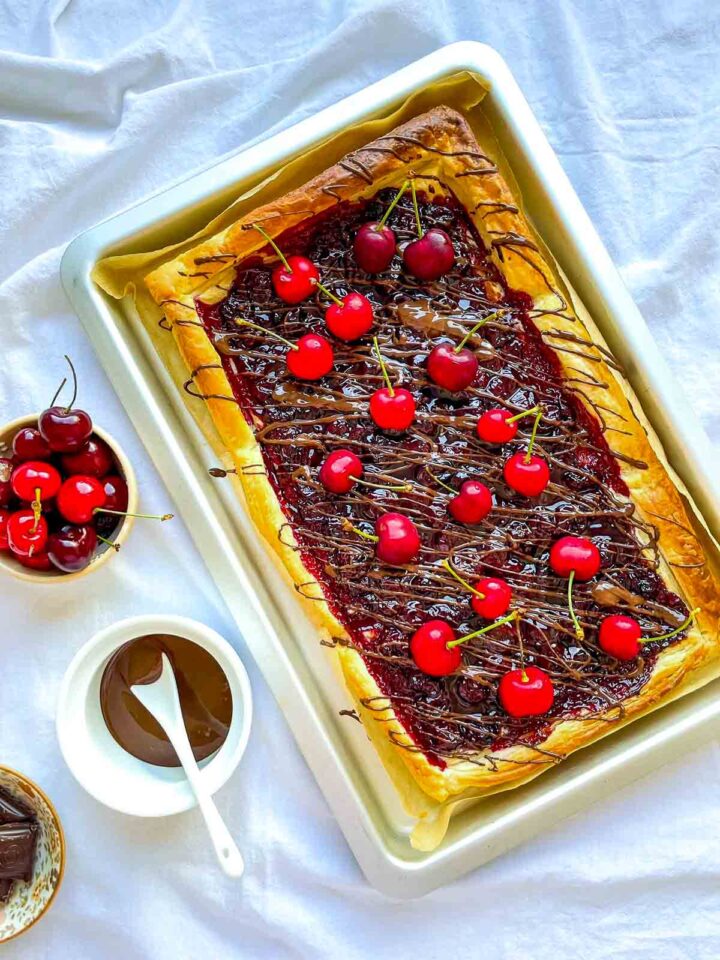 Overhead view of a cherry chocolate puff pastry tart topped with fresh cherries and drizzled chocolate, baked on a tray with bowls of cherries and melted chocolate nearby.