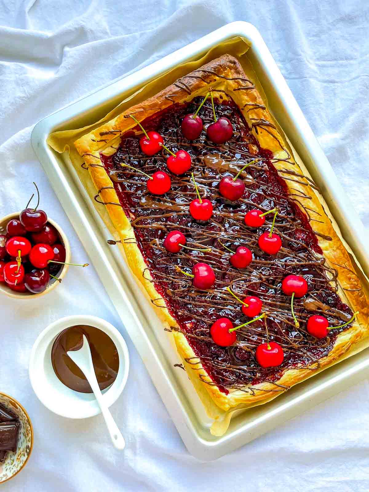 Overhead view of a cherry chocolate puff pastry tart topped with fresh cherries and drizzled chocolate, baked on a tray with bowls of cherries and melted chocolate nearby.