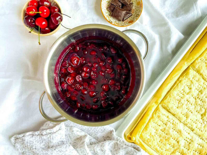 Cherry filling simmering in a saucepan, with fresh cherries, chocolate pieces, and a baked puff pastry crust on a baking tray nearby.