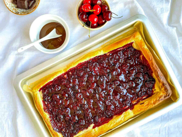 Cherry-filled puff pastry tart drizzled with melted chocolate on a parchment-lined baking tray, with fresh cherries and chocolate sauce nearby.