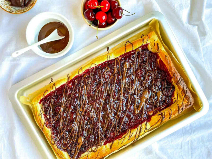 Cherry chocolate puff pastry tart drizzled with melted chocolate on a baking tray, with a bowl of cherries and chocolate sauce nearby.