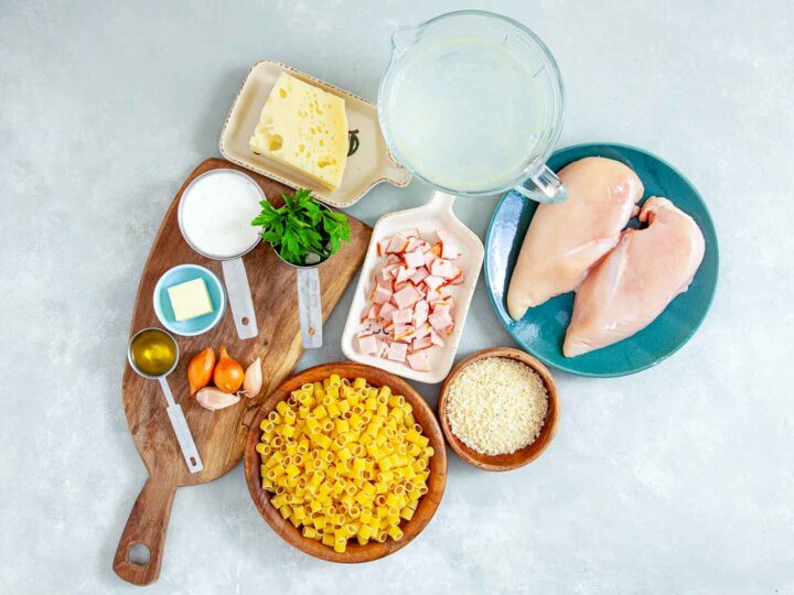 Overhead view of chicken cordon bleu pasta bake ingredients arranged on a light surface, including raw chicken breasts, diced ham, short pasta, shredded cheese, milk, broth, butter, olive oil, garlic, parsley, and breadcrumbs in bowls and measuring cups.
