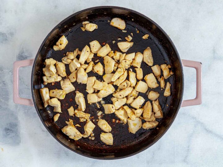 Overhead view of diced chicken breast sautéing in a wide skillet, lightly browned and spaced across the pan as the first step of making chicken cordon bleu pasta bake.