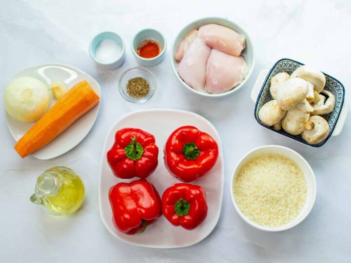 Ingredients for chicken and rice stuffed peppers, including raw chicken, red bell peppers, rice, mushrooms, carrot, onion, spices, and olive oil arranged on a white surface.