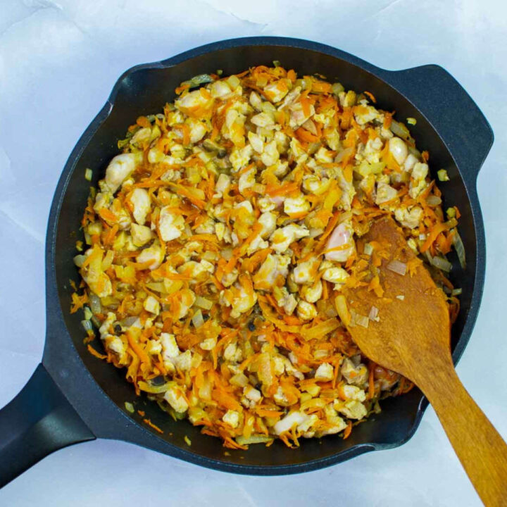 Chicken, rice, and vegetable filling cooking together in a skillet, with chopped chicken, carrots, and onions being stirred with a wooden spoon.