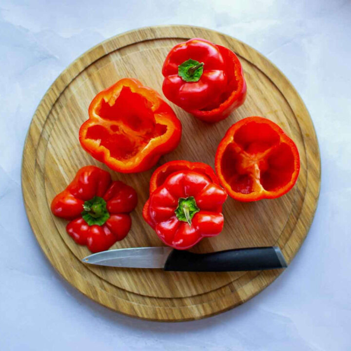 Red bell peppers hollowed and trimmed on a round wooden cutting board, with a small knife beside them, prepared for stuffing.