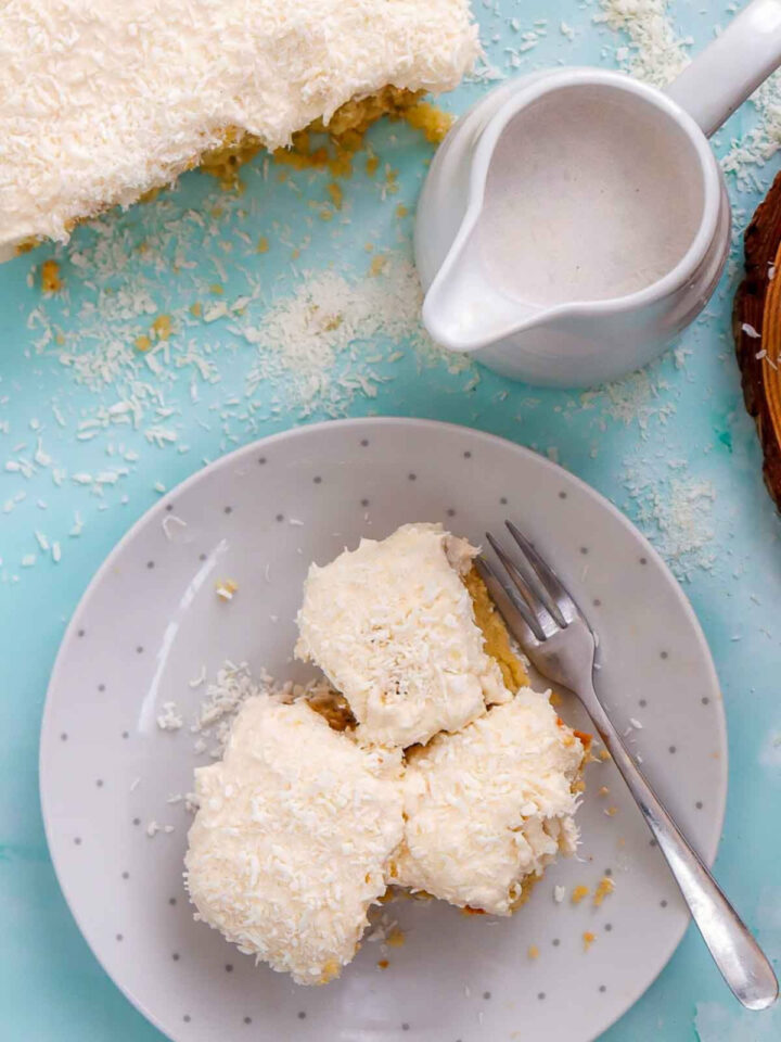 Top-down view of a square slice of coconut cake topped with white frosting and shredded coconut on a polka-dot plate, with a small pitcher of milk and more cake nearby.