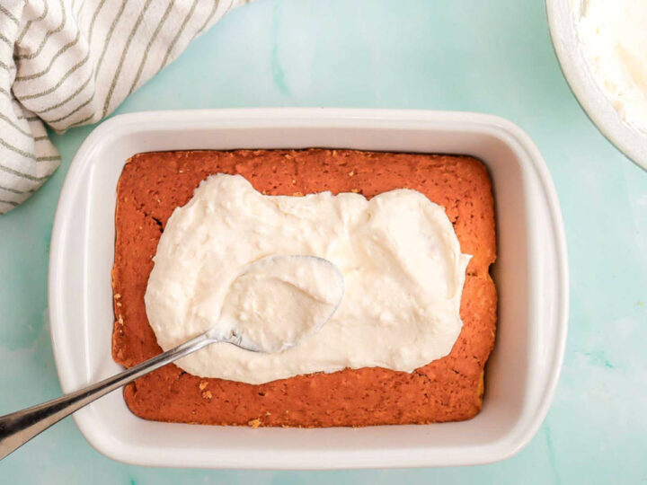 Coconut frosting being spread over a baked coconut cake in a baking dish.