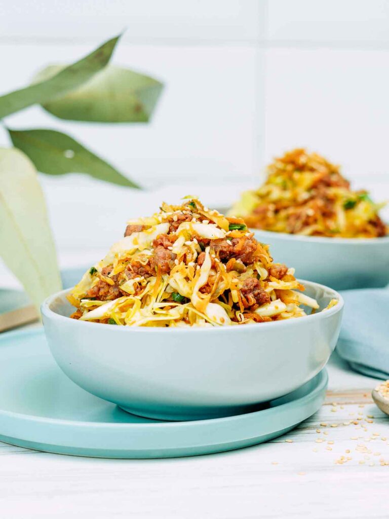 Egg roll bowl with ground meat, shredded cabbage, and vegetables, garnished with sesame seeds and served in a pale blue bowl with a second bowl blurred in the background.