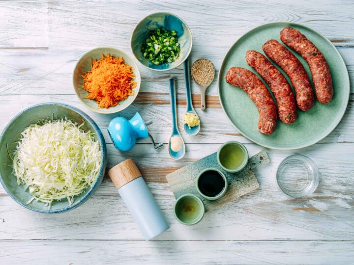 Overhead view of egg roll bowl ingredients arranged on a white wooden surface, including shredded cabbage, grated carrots, chopped green onions, ground sausage links, sesame seeds, minced garlic, soy sauce, oil, and seasonings in small bowls.