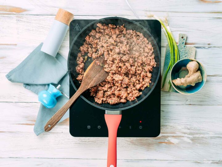 Overhead view of ground meat browning in a skillet on an induction cooktop, with a wooden spatula stirring, steam rising, and fresh ginger and green onions nearby on a light wooden surface.