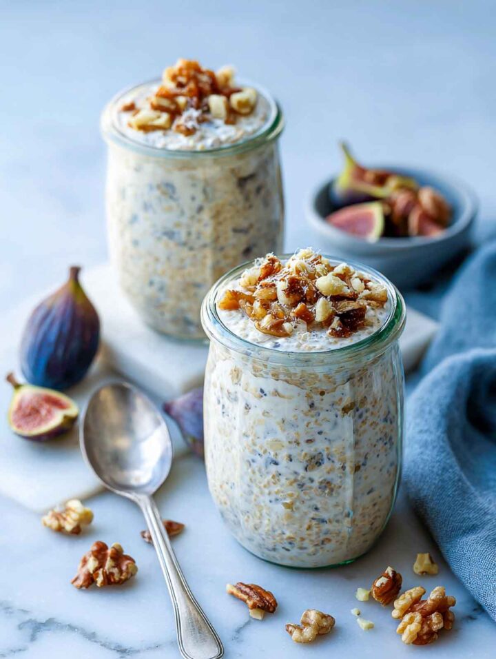 Two glass jars of fig and walnut overnight oats made with steel cut oats, filled close to the rim and topped with chopped dried figs and walnuts, styled on a white marble surface with fresh figs, scattered walnuts, and a blue-gray linen napkin.