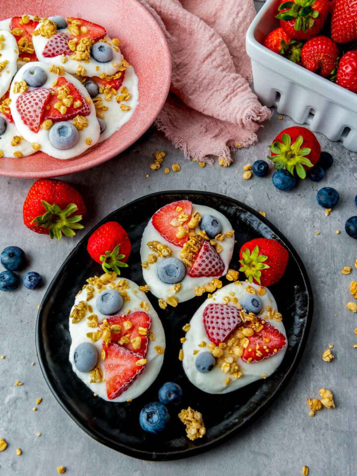 Three dollops of white yogurt topped with sliced strawberries, blueberries, and granola sit on a black plate, surrounded by fresh berries and a container of strawberries on a grey surface.
