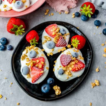 Three dollops of white yogurt topped with sliced strawberries, whole blueberries, and granola sit on a black plate, surrounded by loose berries and granola on a grey surface.