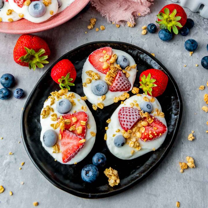 Three dollops of white yogurt topped with sliced strawberries, whole blueberries, and granola sit on a black plate, surrounded by loose berries and granola on a grey surface.