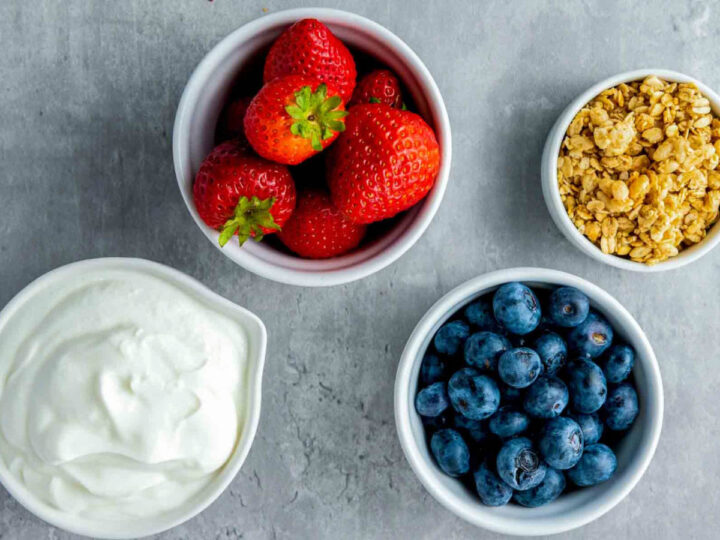 Four small white bowls containing Greek yogurt, fresh strawberries, whole blueberries, and granola are arranged on a grey countertop.