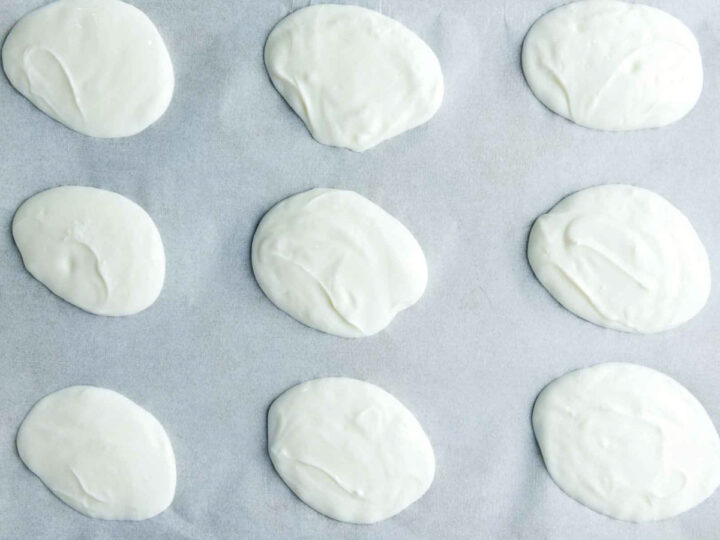 Nine oval-shaped dollops of plain white yogurt are spaced out on a sheet of light grey parchment paper.