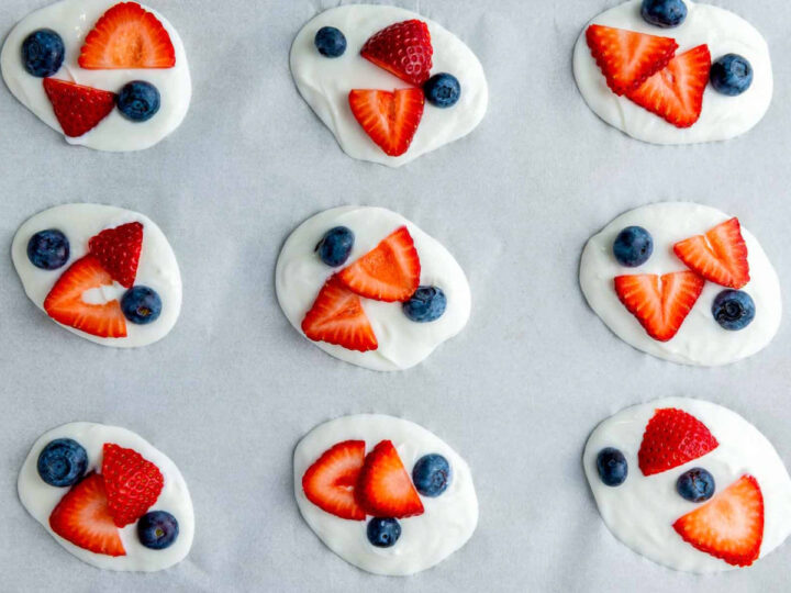 Nine dollops of white yogurt are neatly arranged on a sheet of parchment paper, each topped with sliced strawberries and two whole blueberries.