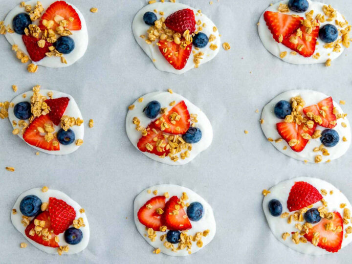 Nine yogurt dollops on parchment paper are topped with sliced strawberries, blueberries, and a final sprinkling of golden granola.