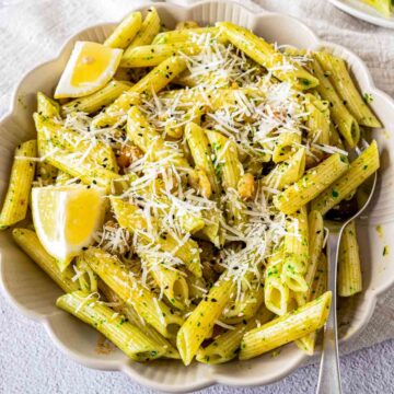 A bowl of penne pasta coated in a light green herb sauce, topped with shredded cheese, small shrimp, and black sesame seeds, garnished with lemon wedges.